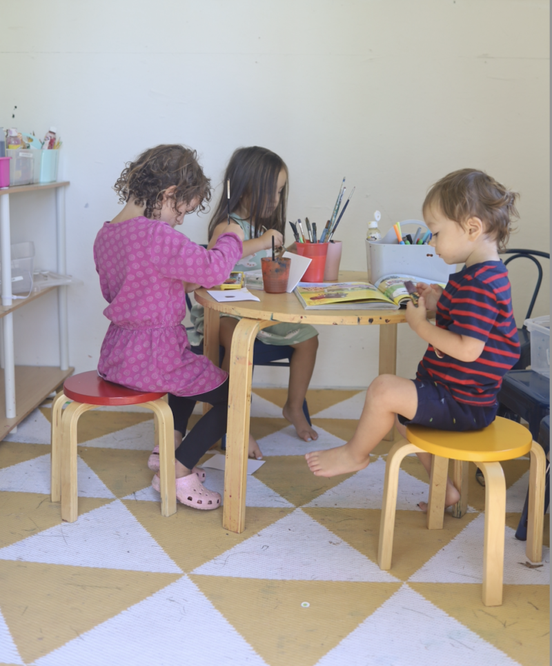 Three children sit at a table looking down at artwork.